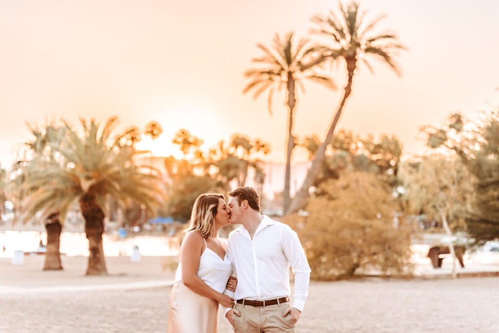 engaged couple kissing with palm trees and sunset behind them